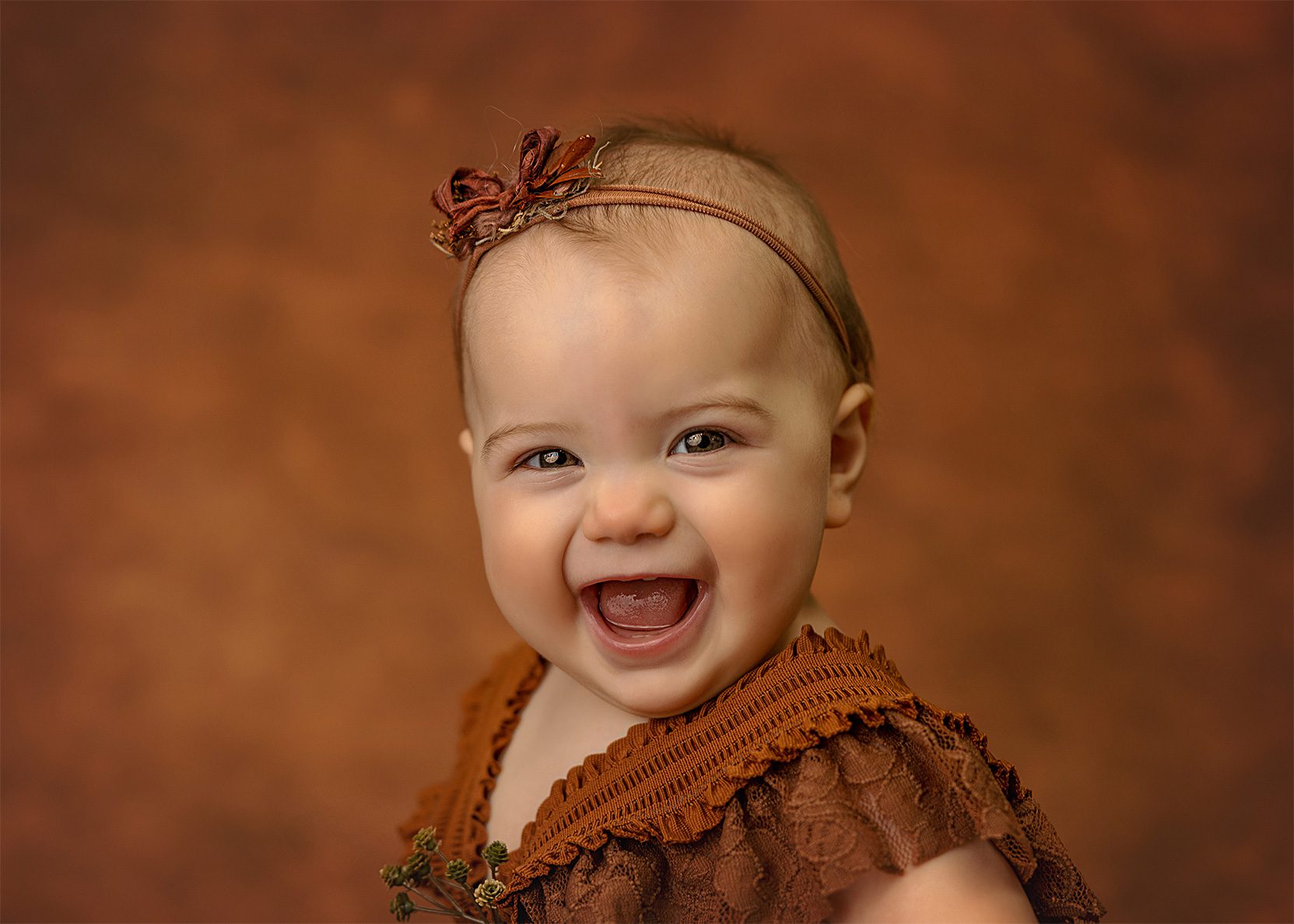 Smiling baby with light skin wearing a brown lace outfit and a brown headband with a bow, set against a soft brown background.