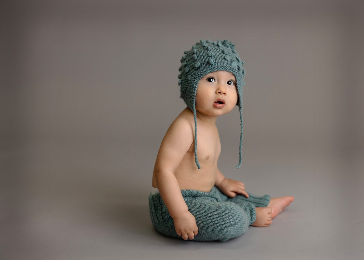 A baby sits on the floor against a plain gray background, wearing a knitted blue-green hat with tassels and matching pants, looking slightly upward with a curious expression.