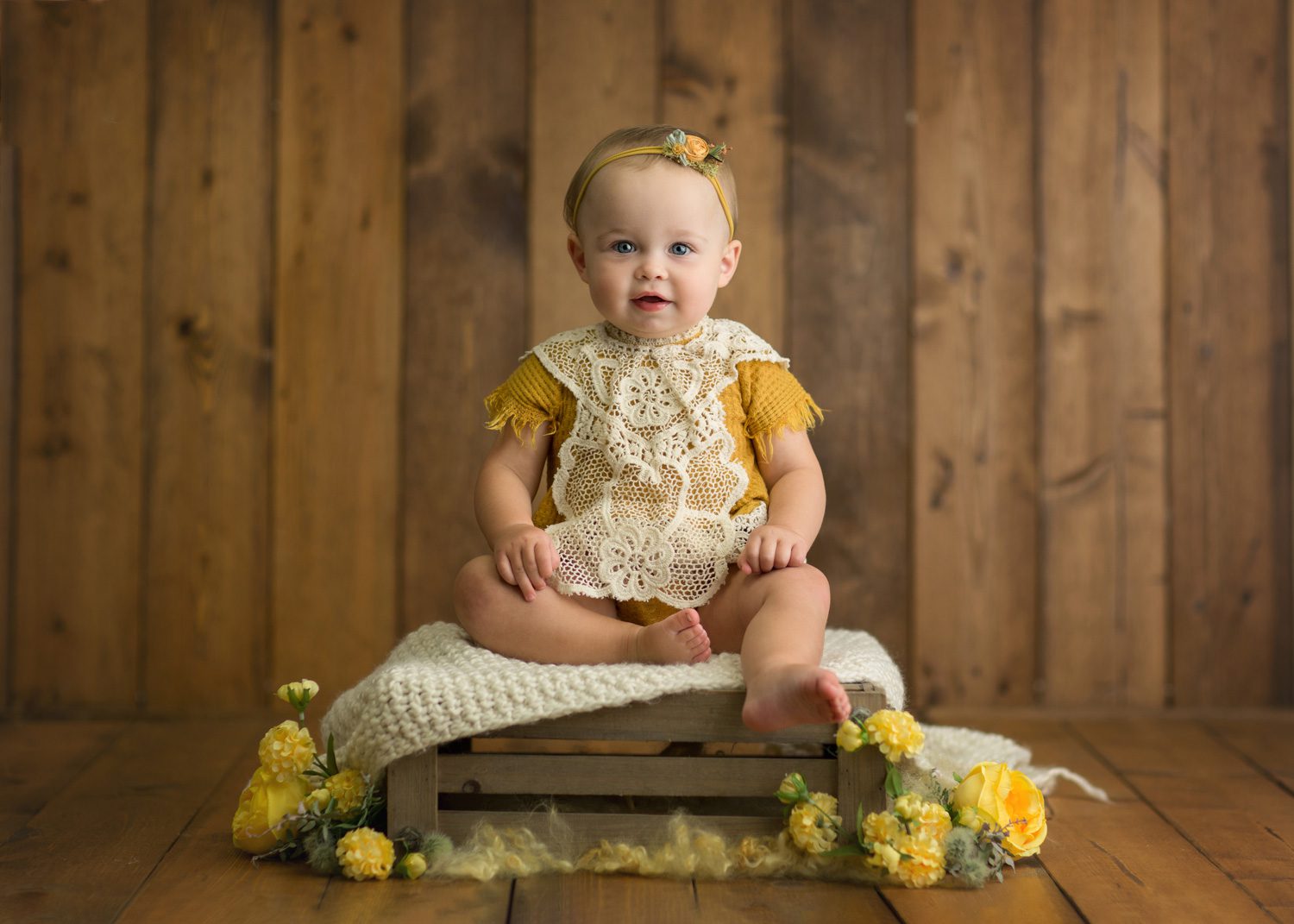 A baby wearing a yellow outfit with a lace bib and headband sits on a wooden crate covered with a knit blanket, surrounded by yellow flowers, against a wooden backdrop.