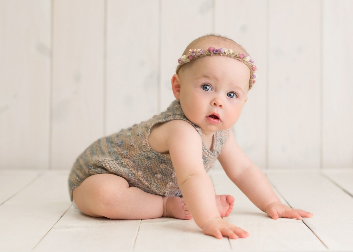 A baby wearing a knit romper and a floral headband sits on a light wooden floor, looking up with a curious expression against a white wood panel background.