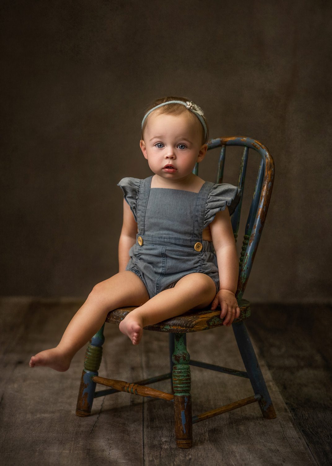 A young child with short hair and a headband sits barefoot on an old wooden chair, wearing a grey romper with ruffled sleeves. The background is brown and softly lit, giving the image a classic, vintage feel.