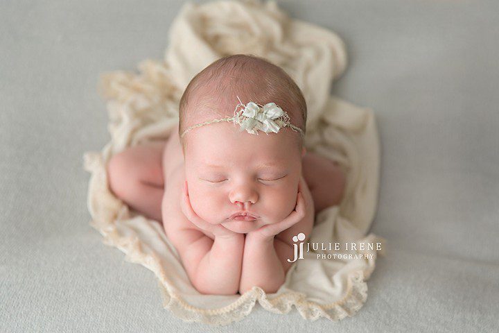 A newborn baby sleeps on a soft cream blanket, with their chin resting on their hands and elbows propped in front. The baby wears a delicate floral headband. Julie Irene Photography logo is visible on the image.