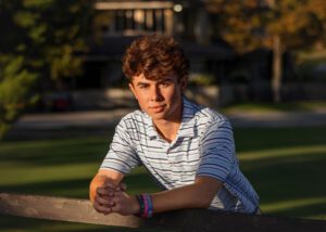 A young man with curly brown hair wearing a striped polo shirt leans on a wooden fence outdoors, with sunlight illuminating his face and a blurred building and greenery in the background.