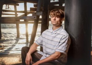 A young man with light brown hair sits against a large pillar under a pier at the beach, wearing a striped button-up shirt, with sunlight shining through the wooden beams and ocean waves in the background.