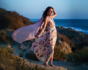 A woman in a flowing floral dress stands barefoot on a rocky beach at sunset, smiling as the wind blows her pink scarf and dress. The ocean and shrubbery are visible in the background.