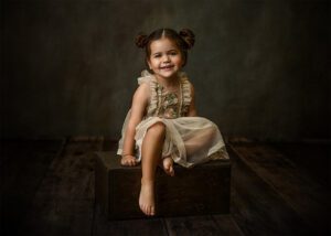A young girl with brown hair in two buns smiles while sitting barefoot on a wooden box, wearing a cream-colored, ruffled dress with floral embroidery against a dark, textured background.