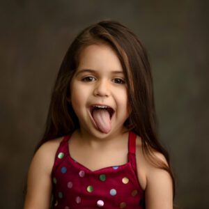 A young girl with long brown hair wearing a sleeveless, red dress with colorful polka dots sticks out her tongue playfully against a muted, dark background.
