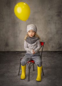 A young child wearing a gray outfit, gray hat, and yellow rain boots sits on a red chair, holding a yellow balloon against a gray backdrop.