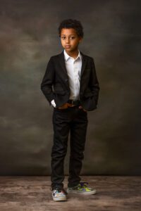 A young boy stands facing forward, wearing a black suit, white dress shirt, and colorful sneakers, with his hands in his pockets against a dark, textured studio background.