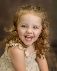 A young girl with curly blonde hair and blue eyes is smiling joyfully at the camera. She is wearing a sleeveless, light-colored dress with ruffled edges, and the background is softly blurred.