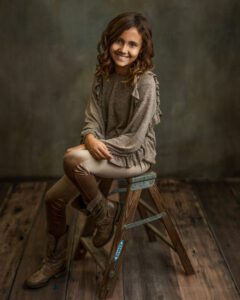 A young girl with wavy brown hair sits on a wooden step stool, smiling at the camera. She wears a taupe sweater, light brown pants, and brown boots, with a rustic wood floor and a muted backdrop behind her.