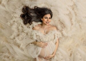 A pregnant woman in a flowing, ruffled cream dress lies on a matching textured background, smiling at the camera with one hand on her belly and long dark hair spread out.