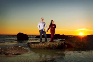 A couple stands on a rock by the ocean at sunset, holding hands. The woman is visibly pregnant and wearing a long red dress, while the man wears a light shirt and jeans. The sky is orange with the sun near the horizon.