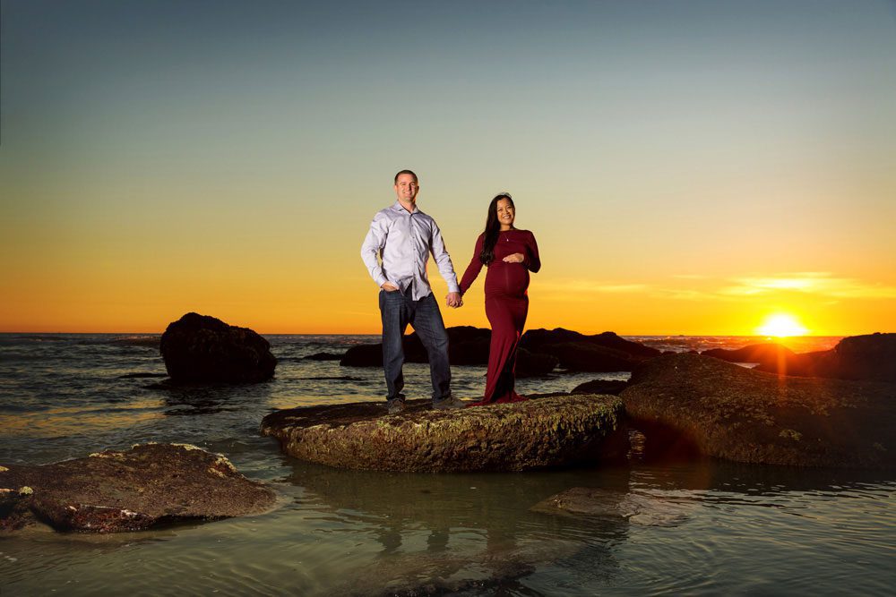 A couple stands on a rock by the ocean at sunset, holding hands. The woman is visibly pregnant and wearing a long red dress, while the man wears a light shirt and jeans. The sky is orange with the sun near the horizon.